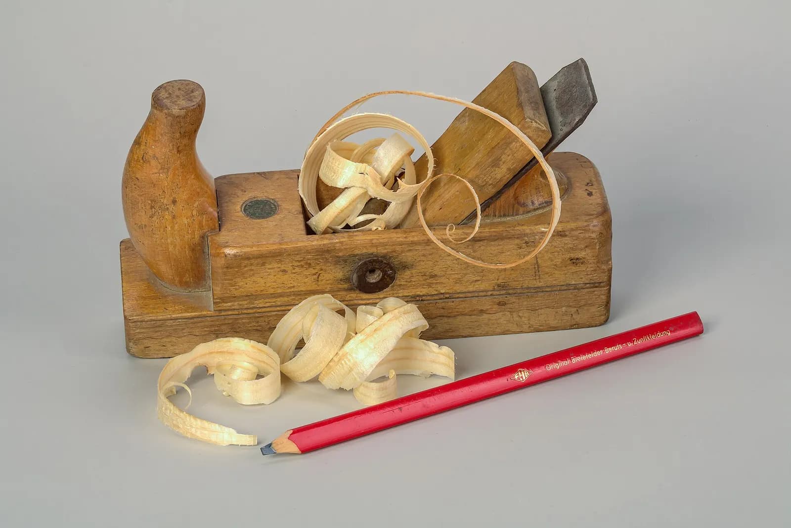 A wooden hand plane, shavings and a red carpenter's pencil.